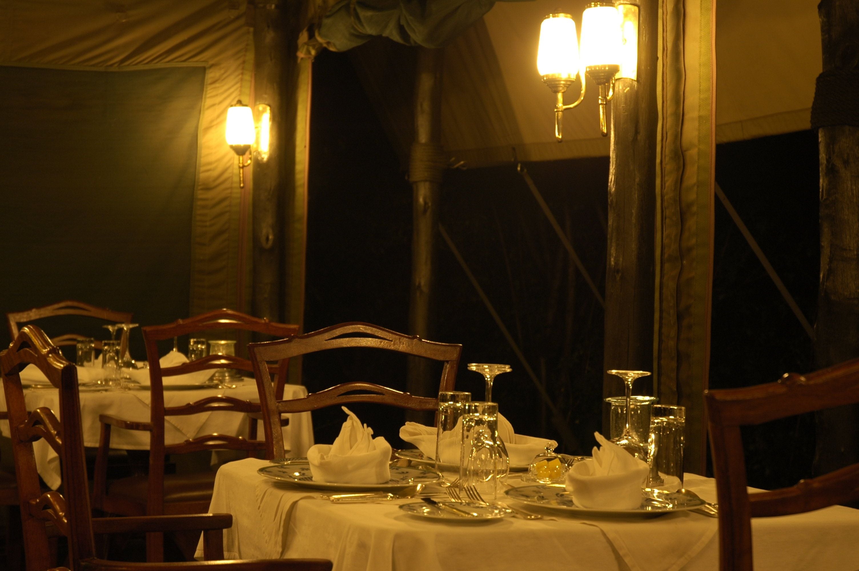 A table set for dining with chairs, a tent-like structure in the background, and wall-mounted lighting.