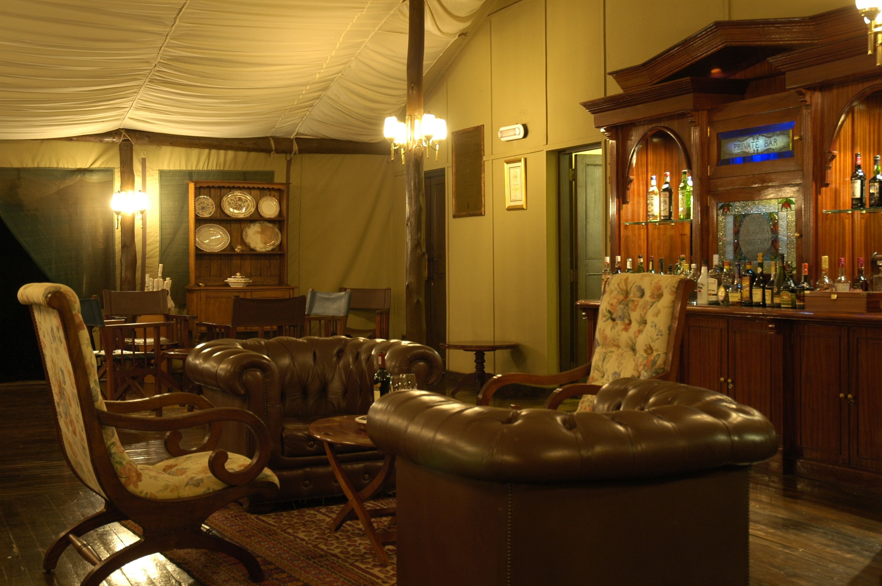 Una zona de bar con mostrador de madera y varias botellas. Hay sillas de cuero y una mesa con una lámpara.