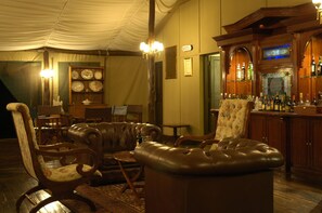 Uma área de bar com balcão de madeira e diversas garrafas. Há poltronas de couro e uma mesa com abajur.