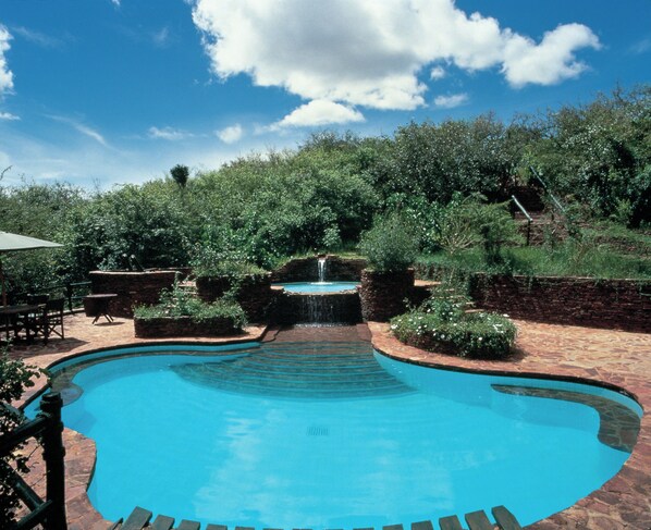 A swimming pool with a waterfall feature, surrounded by greenery and a brick patio.