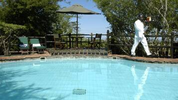 Una piscina con cubierta de madera, sillas de descanso y una persona caminando.