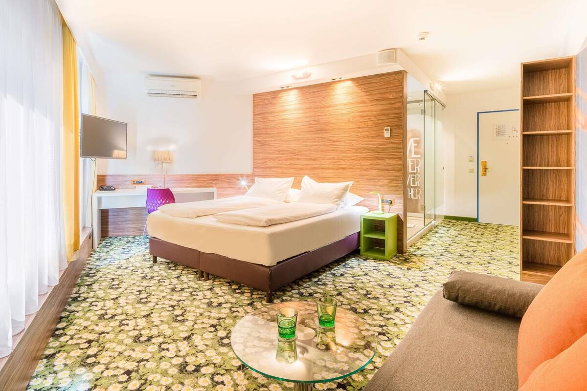 A hotel room with a bed, a glass coffee table with drinks, a sofa, a TV, and a wooden wall.