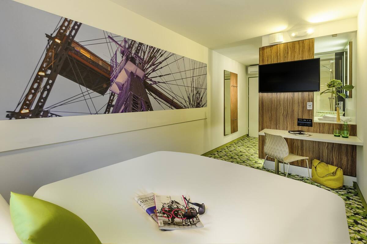 A hotel room with a bed, a desk, a TV, a mirror, and a wall with a Ferris wheel painting.