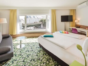 Una camera d'albergo con un letto, una scrivania, una TV e vista su un ponte e palazzi.