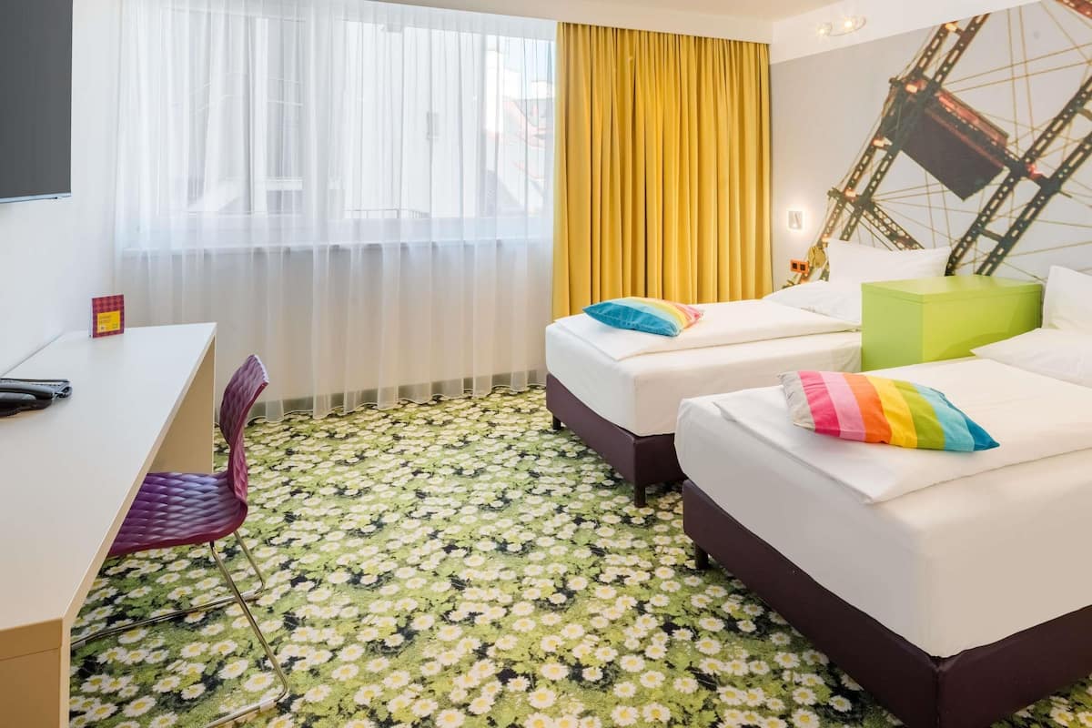 A hotel room with a desk, two beds, a chair, and a large window with curtains.