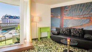 A modern hotel room with a balcony, a large window, a green side table, a black sofa with patterned cushions, and a glass coffee table. - Ibis Styles Wien City (Vienna)