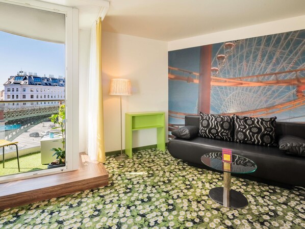 A modern hotel room with a balcony, a large window, a green side table, a black sofa with patterned cushions, and a glass coffee table. - Ibis Styles Wien City (Vienna)