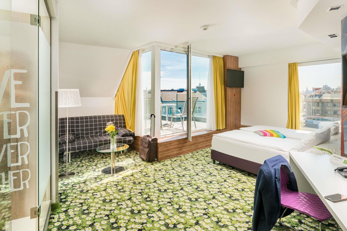 Habitación de hotel con cama, escritorio, silla, televisor y balcón con vistas a la ciudad.
