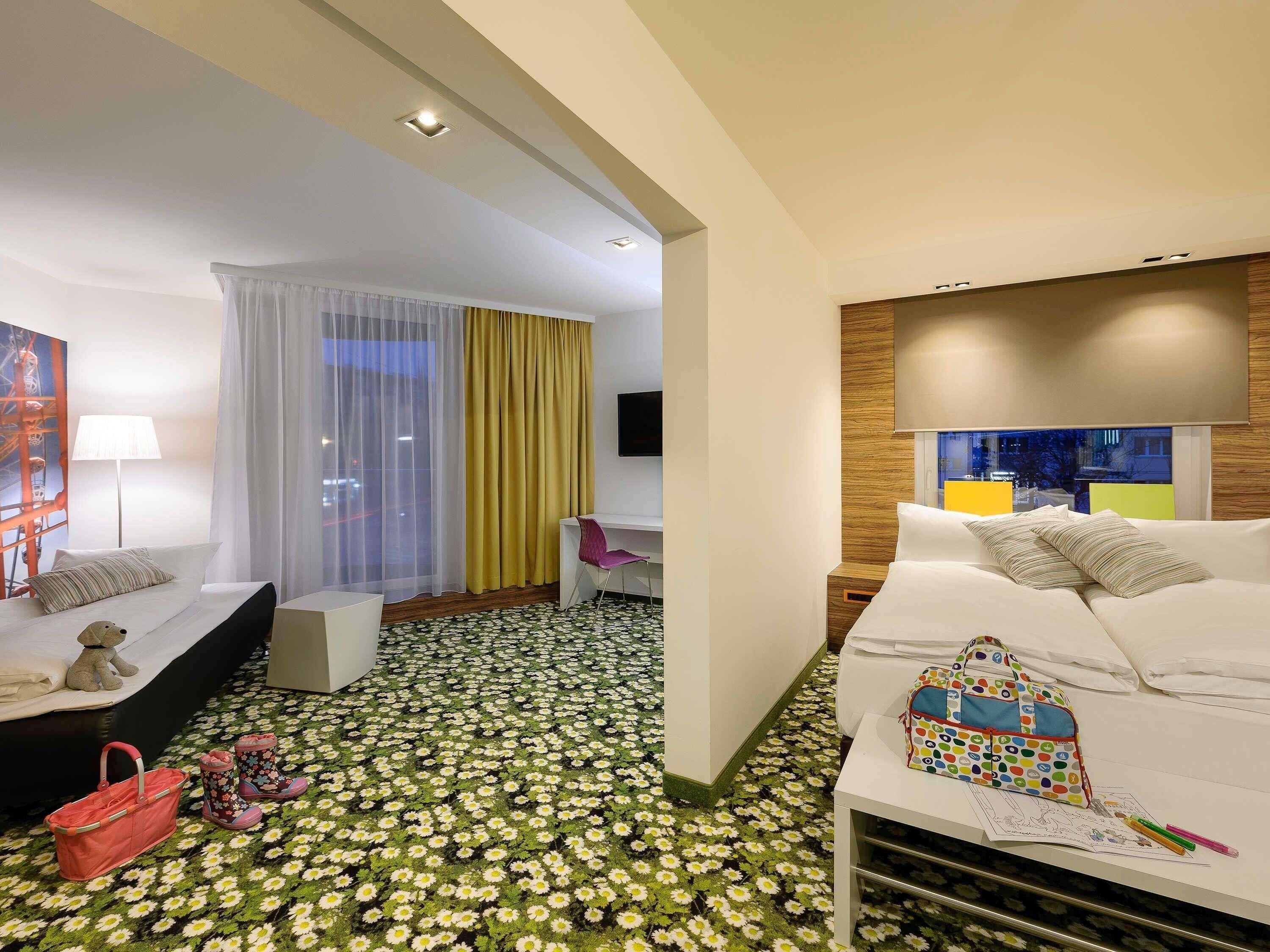 A hotel room with a bed, a desk, a chair, a TV, a window with curtains, and a patterned floor.