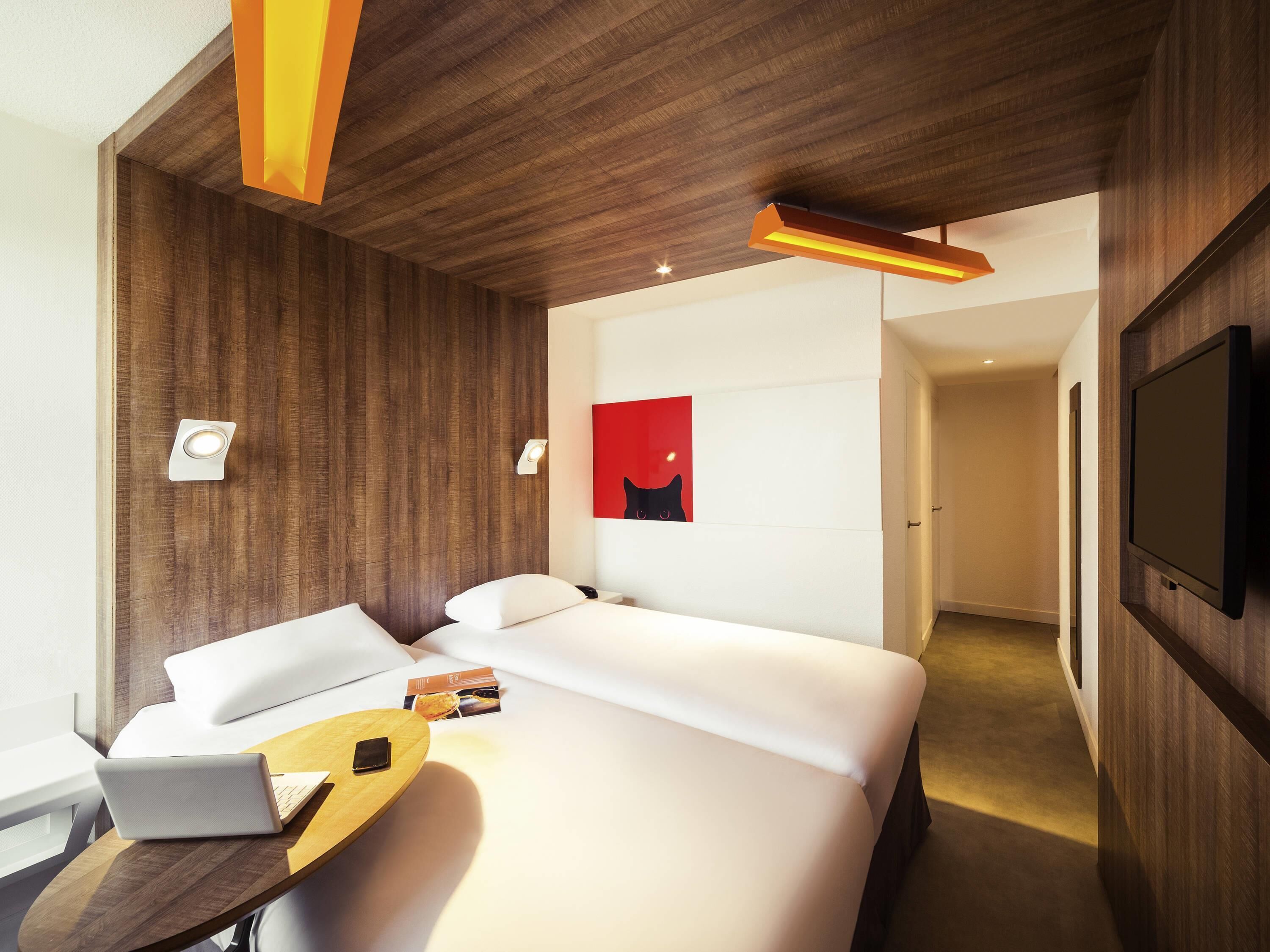 Une chambre d’hôtel moderne avec un grand lit, un bureau avec un ordinateur portable, un téléviseur à écran plat et un tableau abstrait rouge accroché au mur.