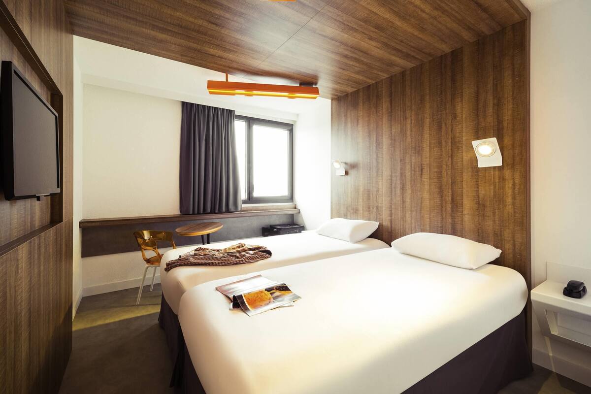 Ein Hotelzimmer mit Bett, Schreibtisch, Stuhl, Fernseher, einem Fenster mit Vorhängen und einer Holzbalkendecke.