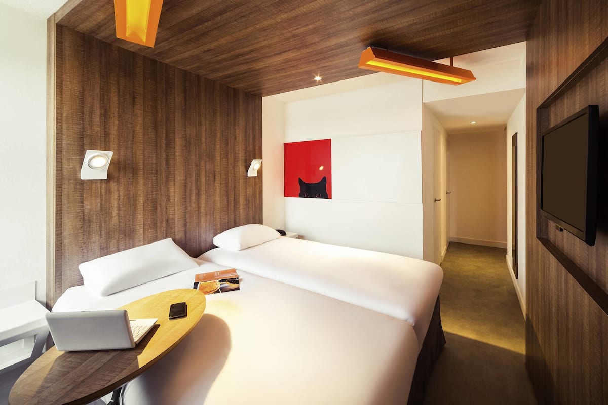 A modern hotel room with a large bed, a desk with a laptop, a flat-screen TV, and a red abstract painting on the wall.