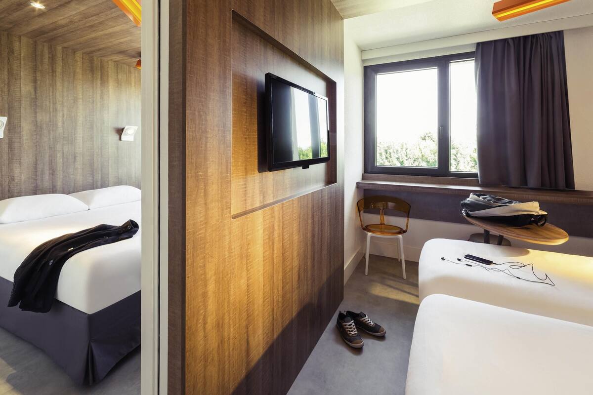 Ein Hotelzimmer mit einer Wand in Holzoptik, einem Flachbildfernseher, einem Fenster mit Vorhängen, einem Sessel, einem kleinen Tisch mit einer Tasche und einem Paar Schuhen auf dem Boden.