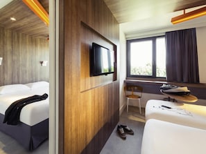 A hotel room with a wooden accent wall, a flat-screen TV, a window with curtains, a chair, a small table with a bag, and a pair of shoes on the floor.