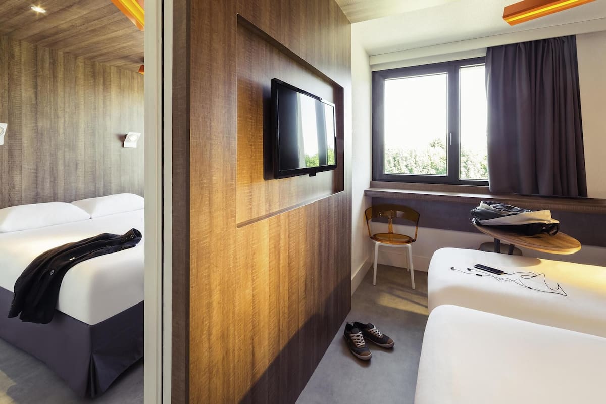 A hotel room with two beds, a TV, a window with curtains, a chair, a table, and a pair of shoes.