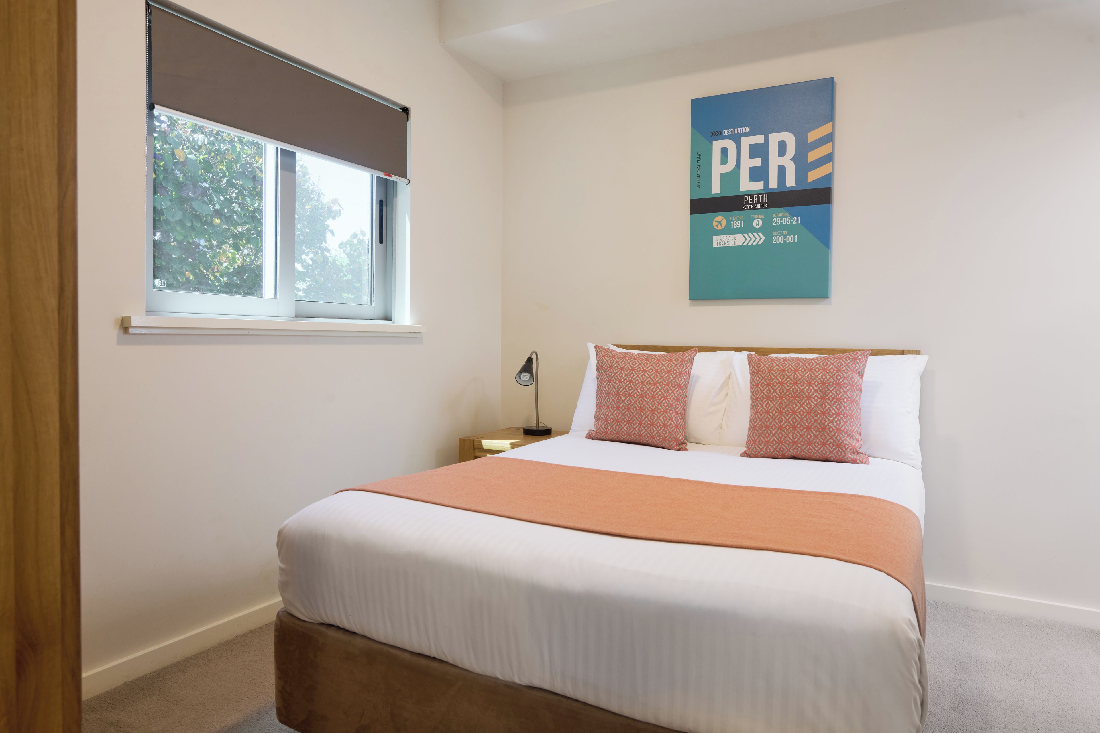 A hotel room with a bed, a window with a blind, a nightstand, and a wall-mounted sign.