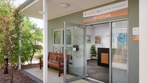 An airport reception area with a wooden bench, glass doors, and a sign for VetroBlu Airport Apartments. - Airport Apartments by Vetroblu (Redcliffe)