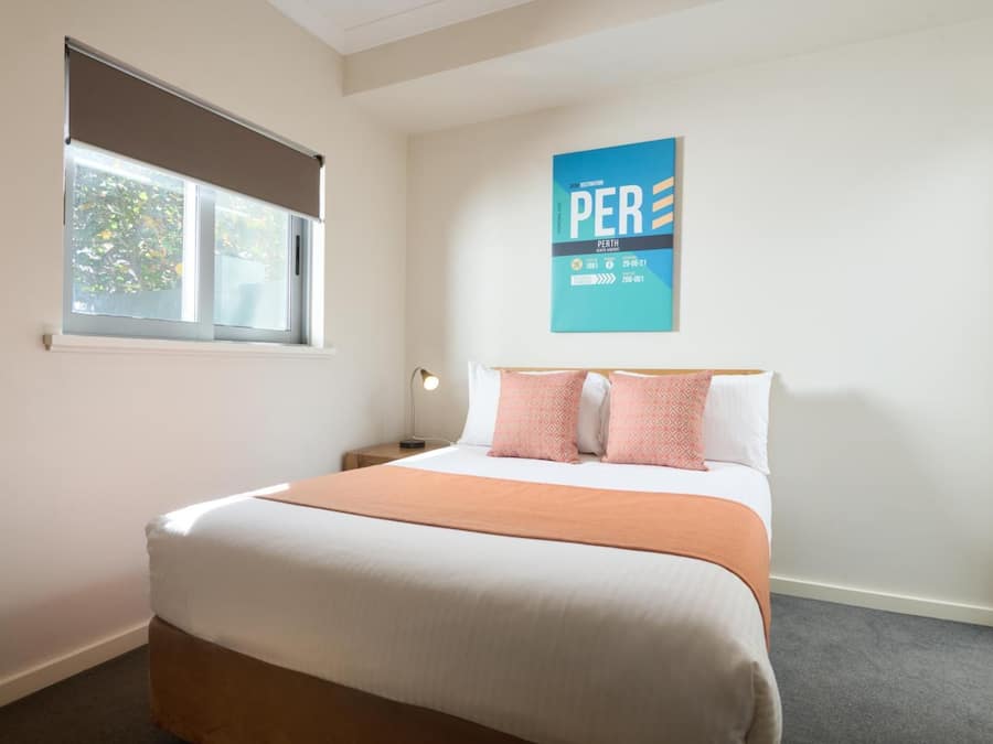 A hotel room with a bed, a window with blinds, a nightstand, a lamp, and a wall poster.