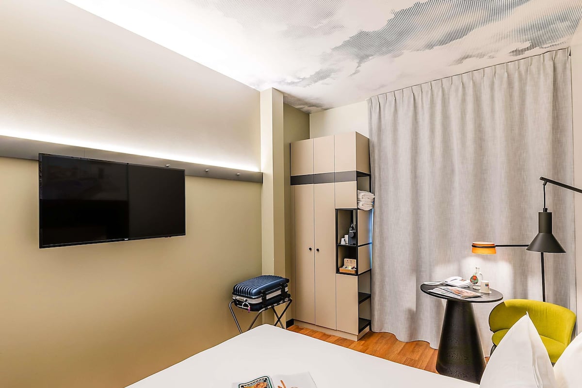A hotel room with a flat-screen TV mounted on the wall, a bed with a magazine, a small table with a lamp, a chair, and a wardrobe.