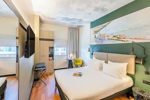 A hotel room with a bed, a chair, a desk, a television, and a painting on the wall. - ibis Lisboa Parque das Naçoes (Lisbon)