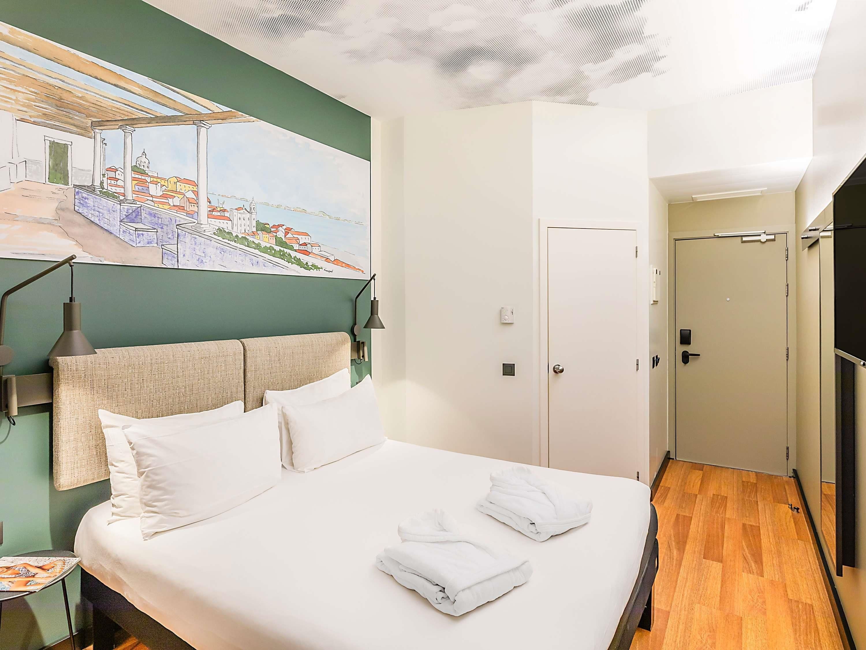 A hotel room with a bed, a nightstand, a painting of a cityscape, a door, and a wooden floor.
