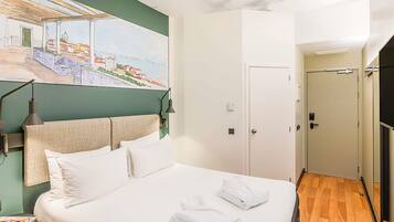 A hotel room with a bed, a nightstand, a painting of a cityscape, a door, and a wooden floor.