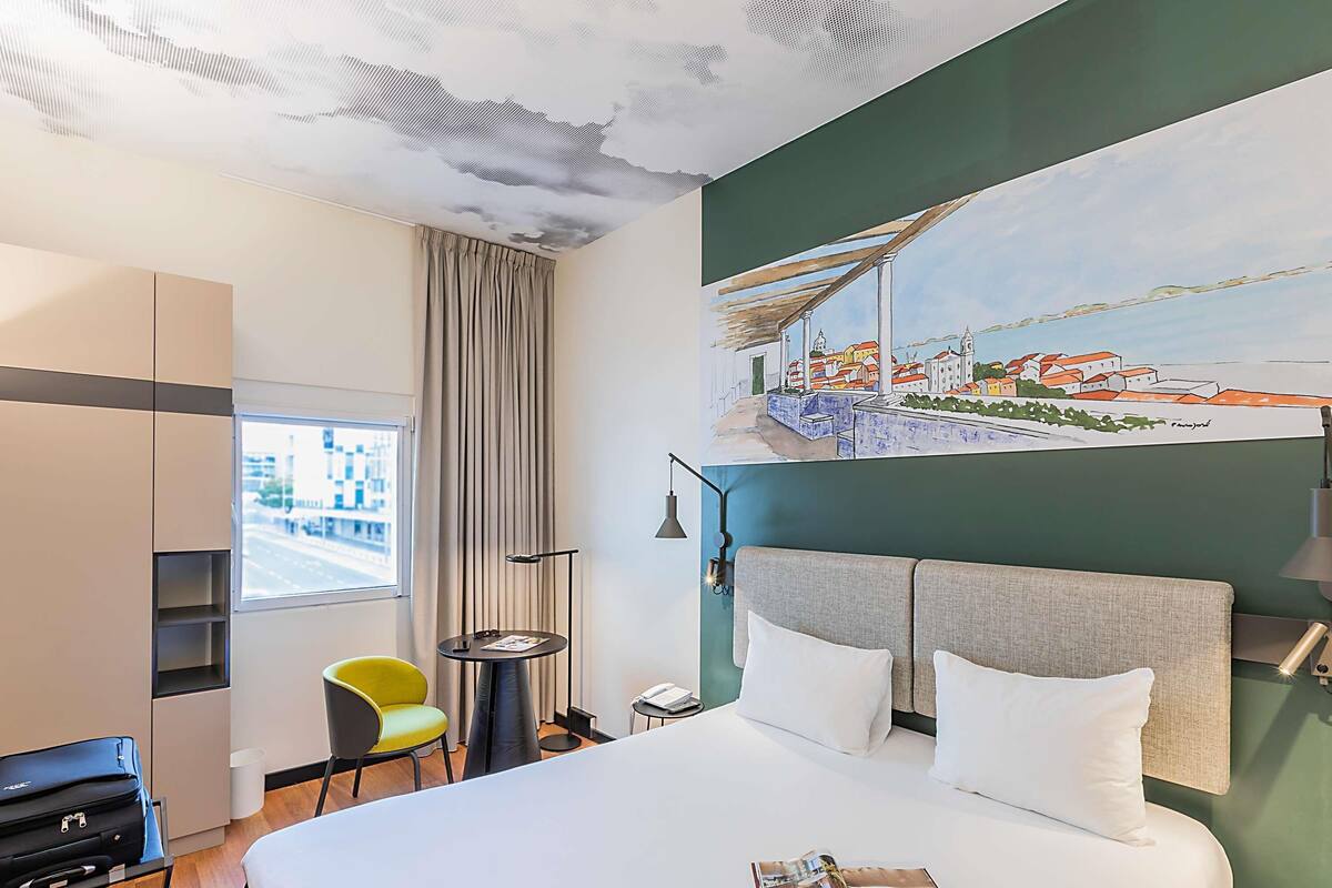 A hotel room with a large bed, a view of the city through the window, a painting on the wall, and a small table with a lamp.