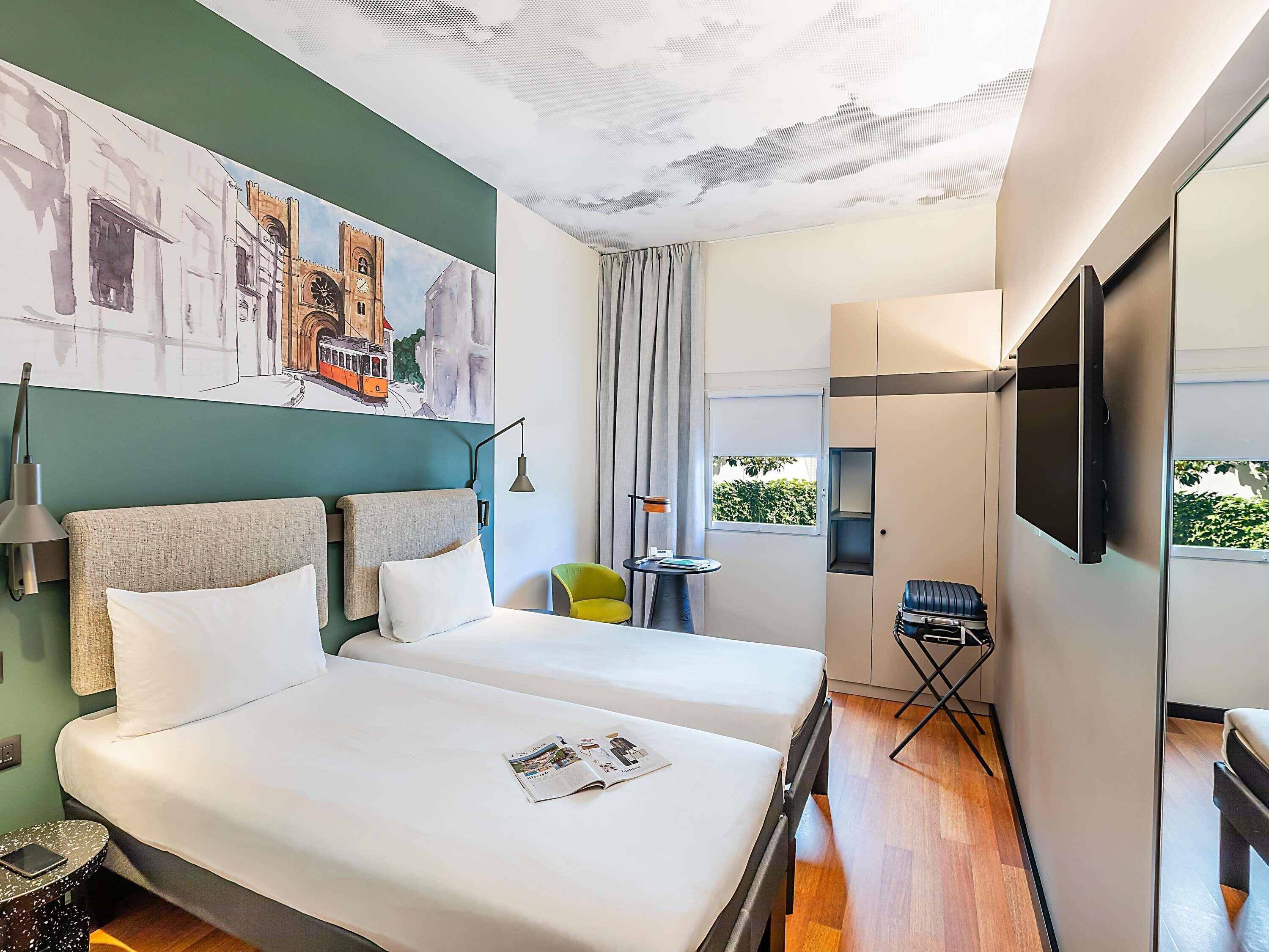A hotel room with two beds, a TV, a window with a view, a painting on the wall, and a small table with a chair.