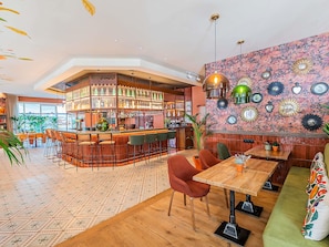 Zona de bar con mostrador curvo, varios taburetes y una amplia variedad de botellas y vasos. Hay mesas y sillas para sentarse, y las paredes están decoradas con espejos y obras de arte.