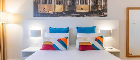Ein Hotelzimmer mit einem Bett, zwei Nachttischen, einer Lampe, einem Gemälde mit Straßenbahnen und einer blauen Decke.