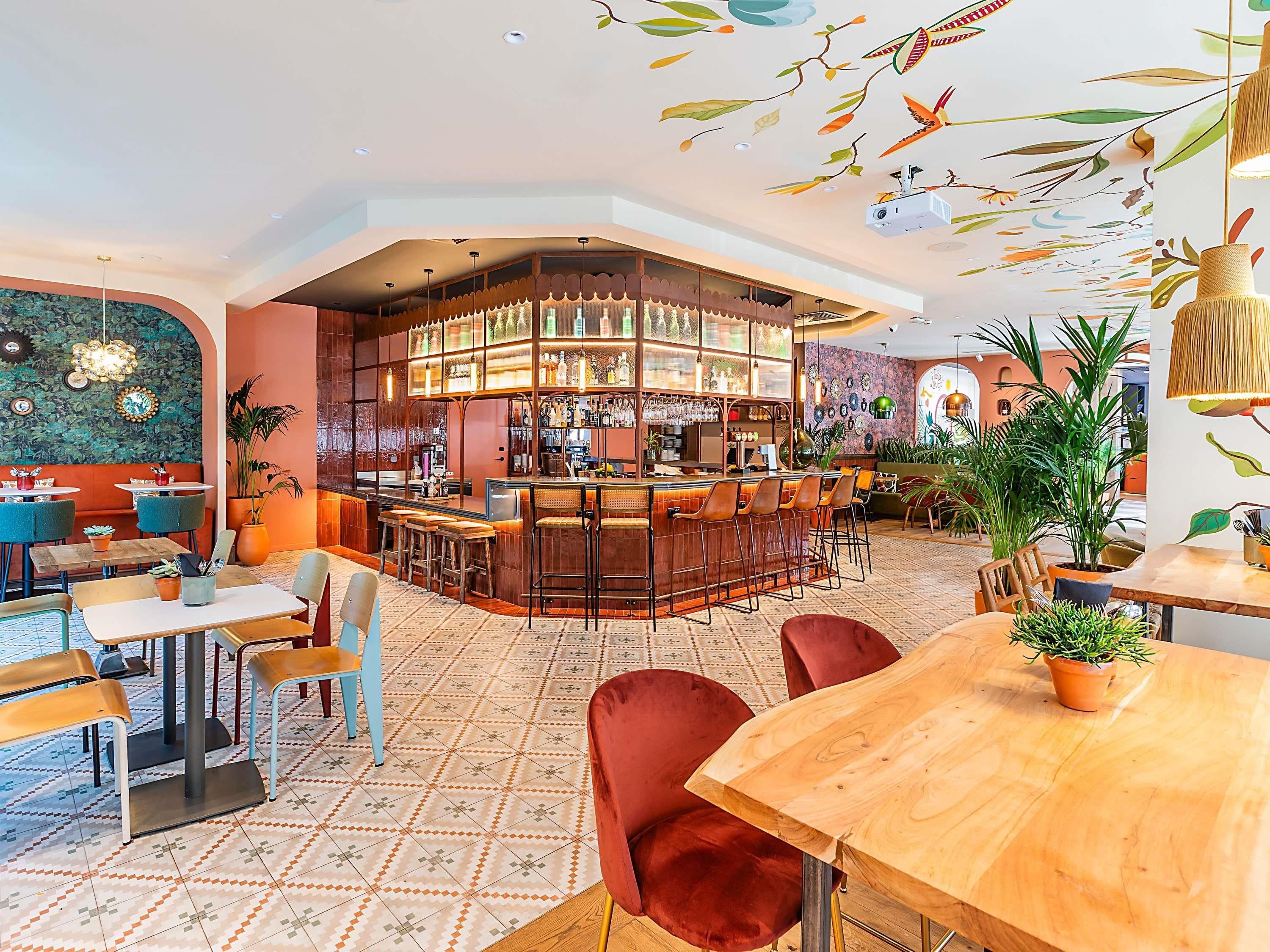 Uma área de bar com balcão, banquetas e diversos frascos e copos. Há mesas e cadeiras ao redor do bar, e o chão possui um desenho de azulejos padronizados.