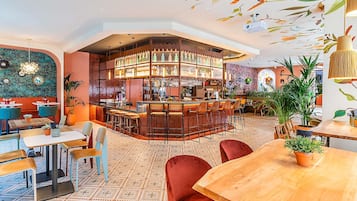 Uma área de bar com balcão, banquetas e diversos frascos e copos. Há mesas e cadeiras ao redor do bar, e o chão possui um desenho de azulejos padronizados.