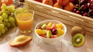Een ontbijttafel met een kom gemengd fruit, een glas sinaasappelsap en diverse verse vruchten zoals druiven, kersen en kiwi.