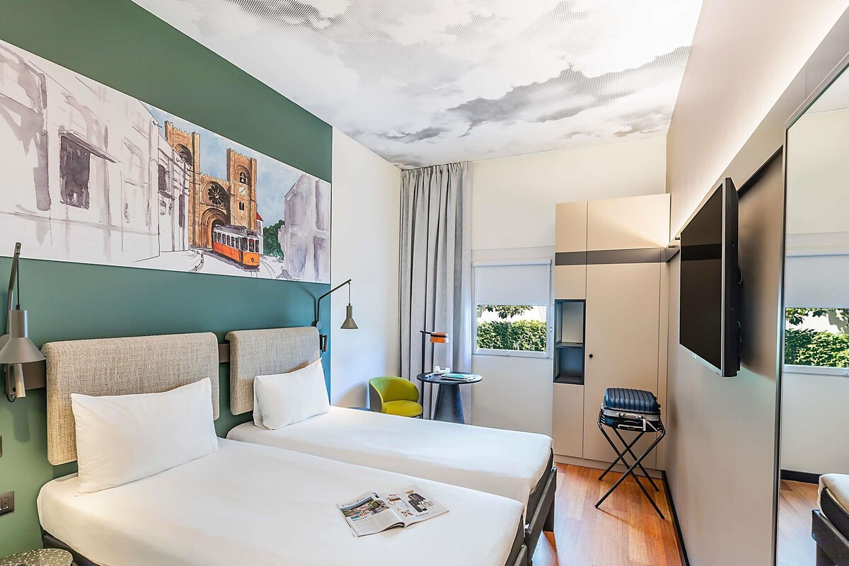 A hotel room with two beds, a TV, a window with a view, a painting on the wall, and a small table with a chair.