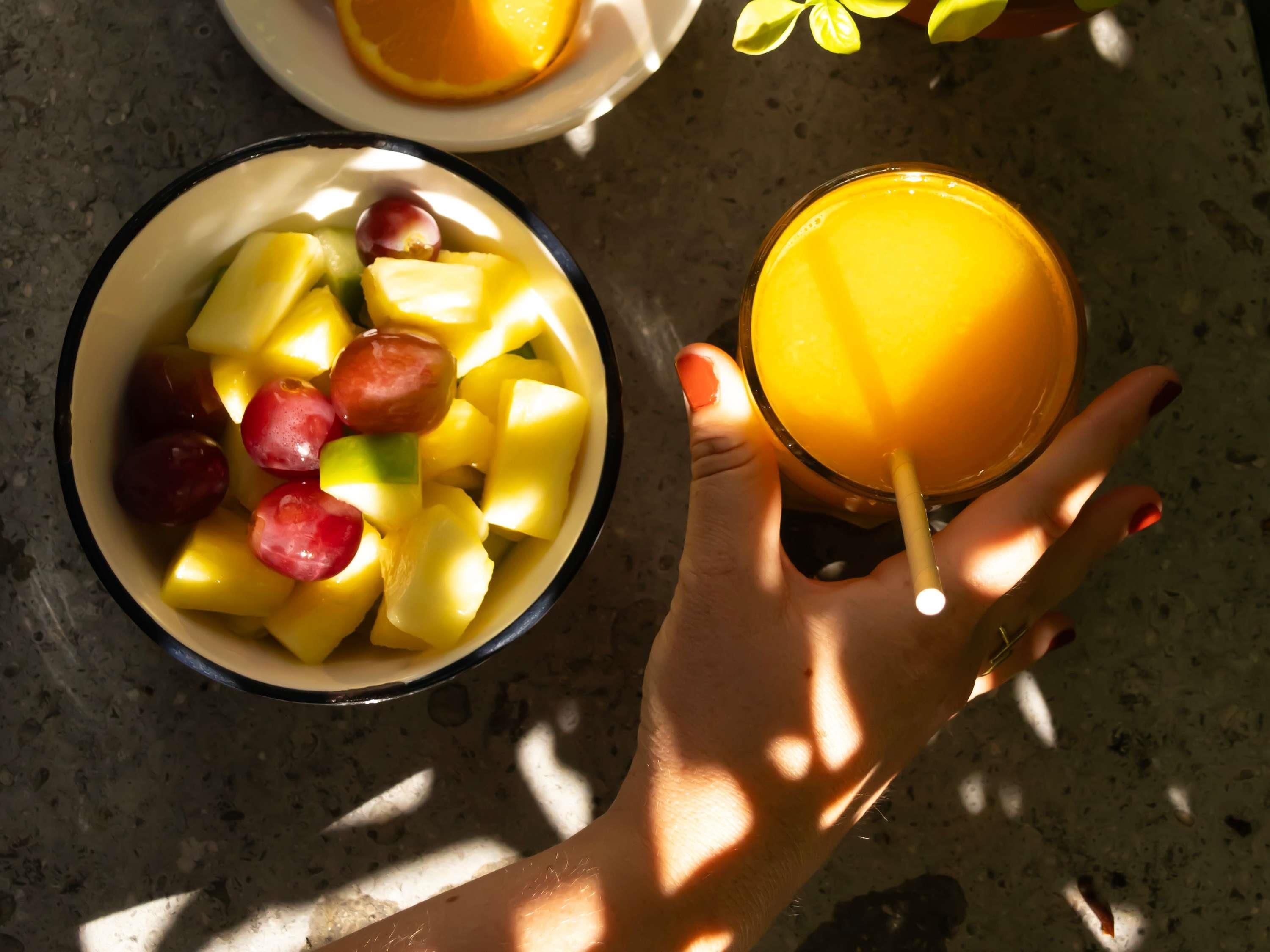 Een hand die een glas sinaasappelsap vasthoudt, naast een schaal fruit.