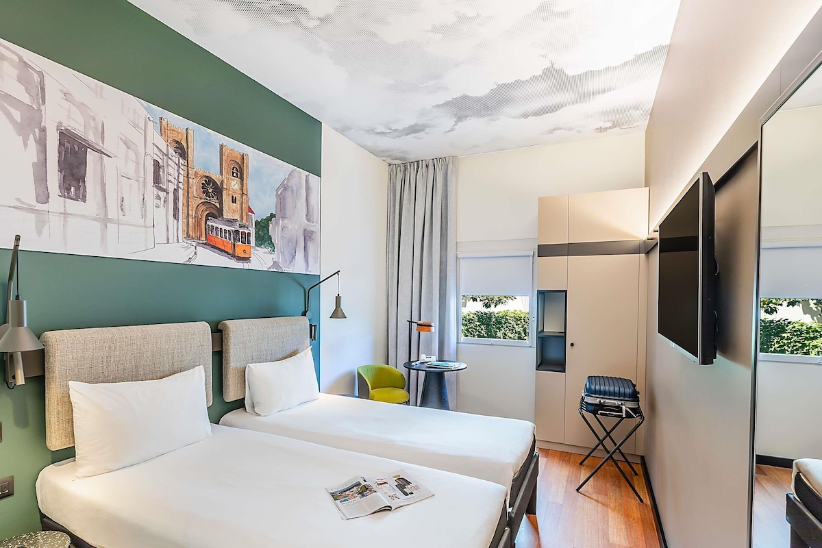 Une chambre d’hôtel avec deux lits, une télévision, une fenêtre donnant sur un beau paysage, un tableau au mur et une petite table avec une chaise.