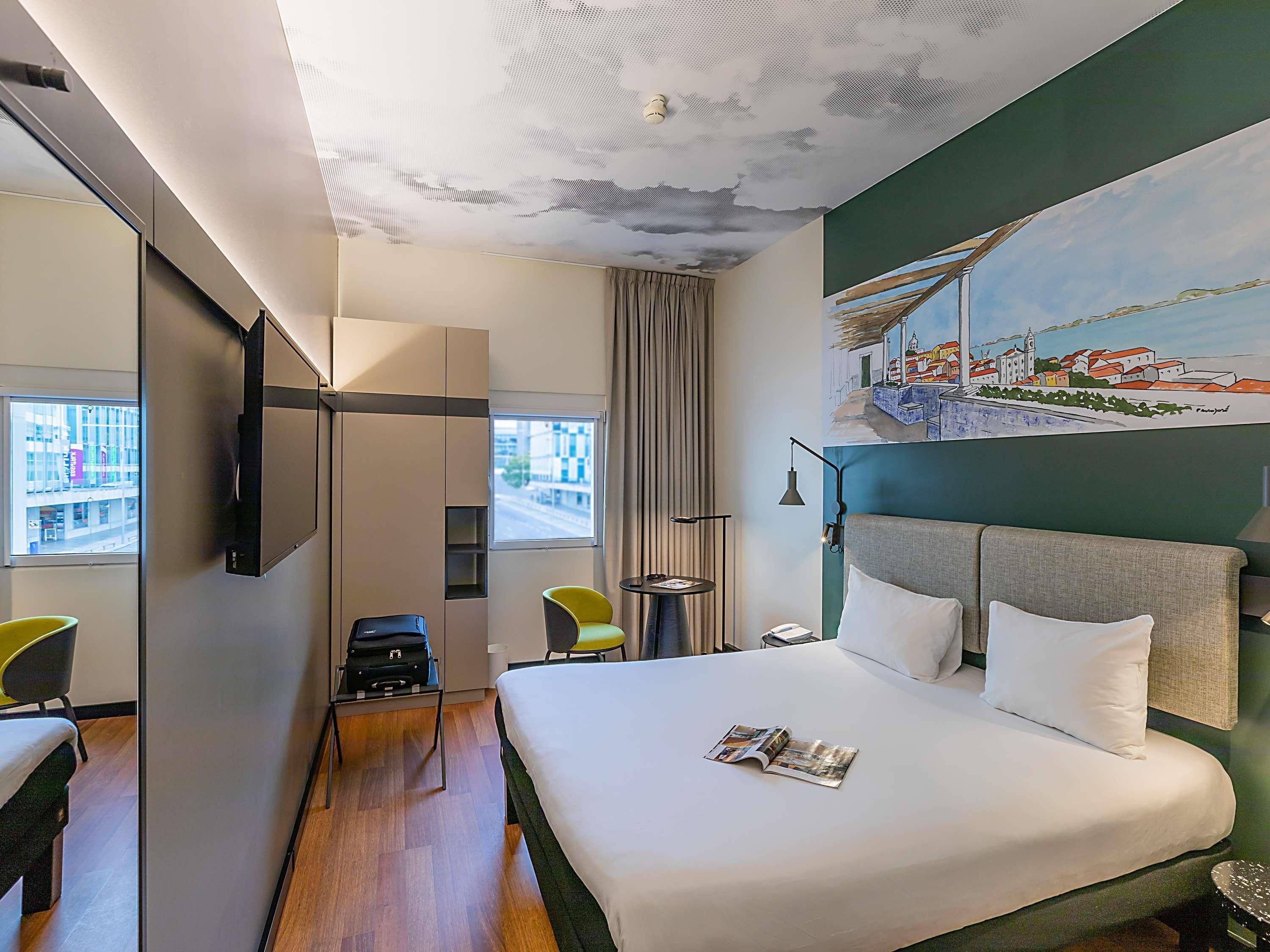 A hotel room with a bed, a desk, a chair, a TV, and a painting on the wall.
