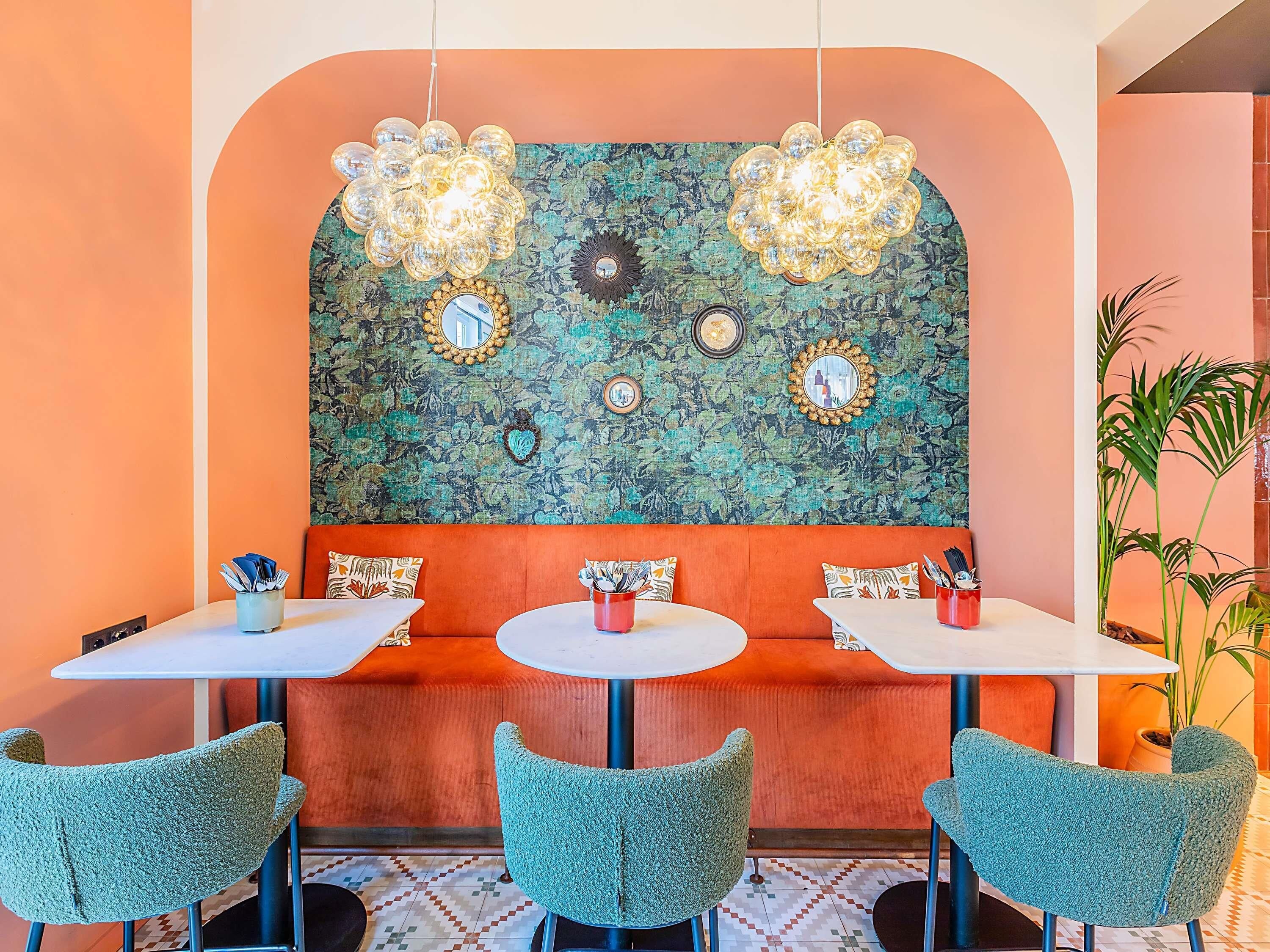 A restaurant with a vibrant orange wall, a green floral mural, and a seating area with teal chairs and orange cushioned benches.
