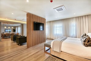 A modern hotel room with a large bed, a TV mounted on a wooden panel, a dining area in the background, and a view of the outdoors through large windows.