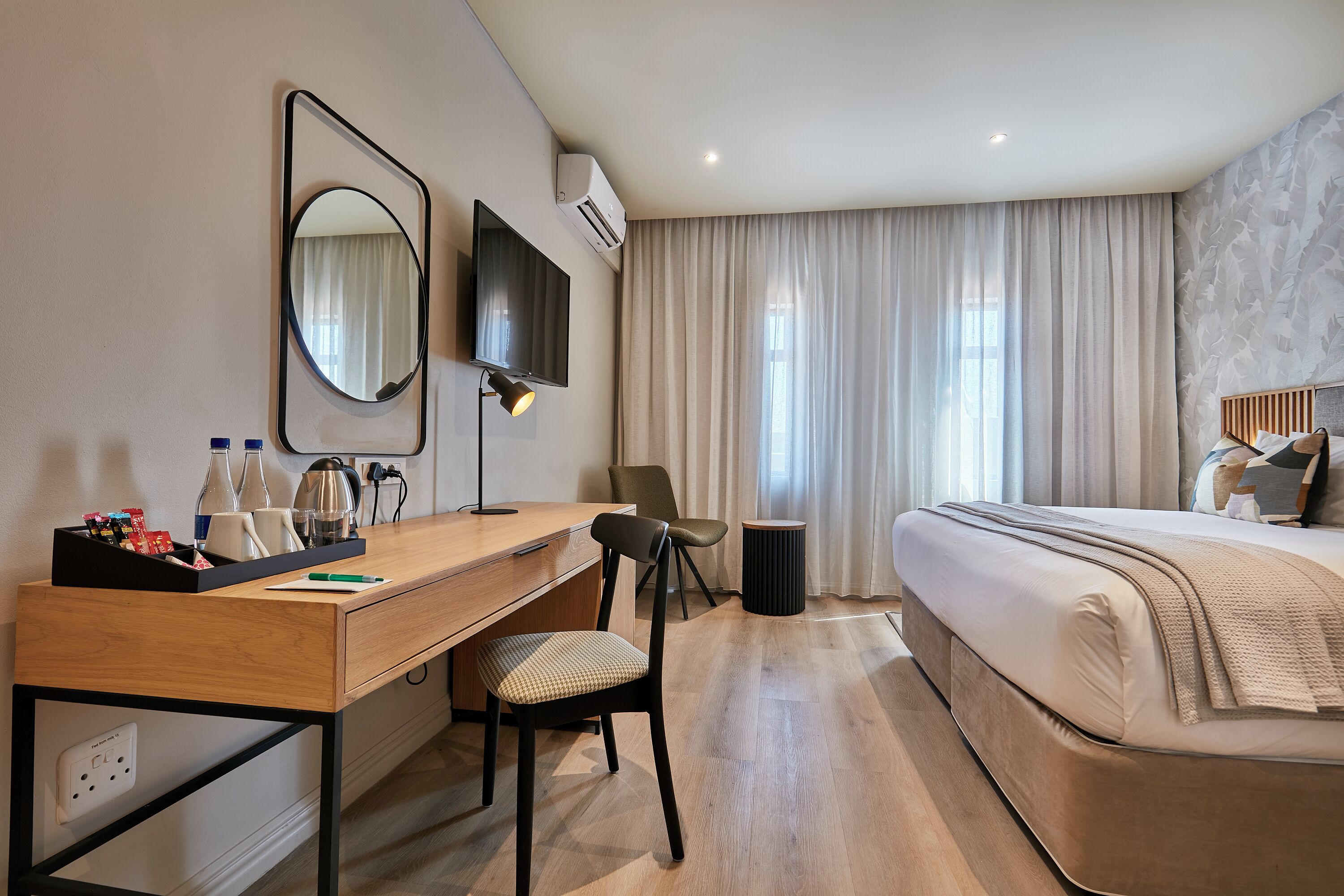 A hotel room with a wooden desk, a chair, a bed with a beige comforter, a TV mounted on the wall, and a window with curtains.