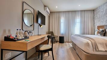 A hotel room with a wooden desk, a chair, a bed with a beige comforter, a TV mounted on the wall, and a window with curtains.