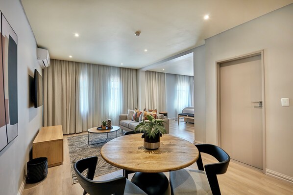 A modern living room with a round wooden table, black chairs, a sofa, and a large window with curtains.