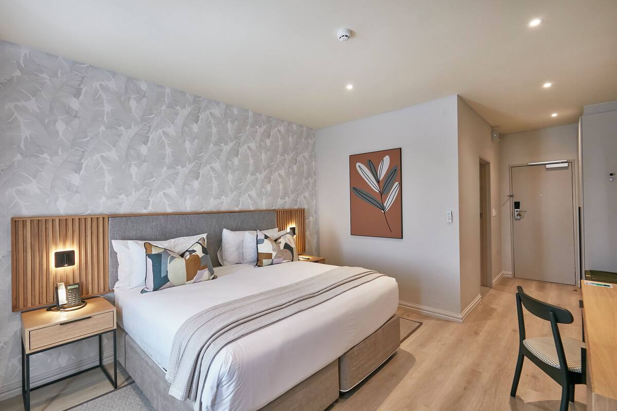 A modern hotel room with a large bed, bedside lamps, a wooden headboard, a wall-mounted artwork, and a door leading to a bathroom.