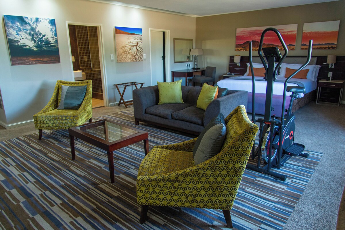 A hotel room with a bed, exercise equipment, a sofa, two armchairs, a coffee table, and artwork on the walls.
