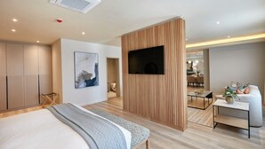 A modern bedroom with a bed, a TV mounted on a wooden panel, a seating area with a table, and a view into the kitchen.