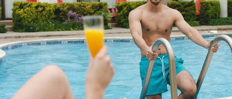 Escena junto a la piscina con una persona en bañador azul de pie junto a ella, sosteniendo una escalera de piscina. Otra persona se relaja en una tumbona, sosteniendo un vaso de jugo de naranja.