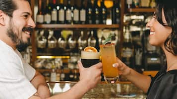 Dos personas brindando con bebidas en un bar, con estantes llenos de botellas al fondo.