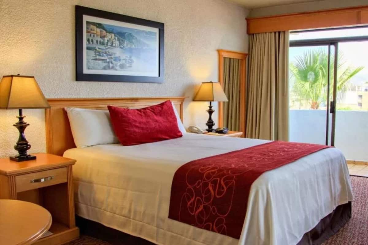 A hotel room with a bed, two bedside lamps, a painting on the wall, a window with curtains, and an air conditioning unit.