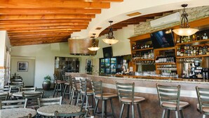 A bar area with wooden ceiling beams, a stone wall, and a bar counter with various bottles and glasses. There are multiple wooden chairs and tables around the bar. - Corona Hotel & Spa (Ensenada)