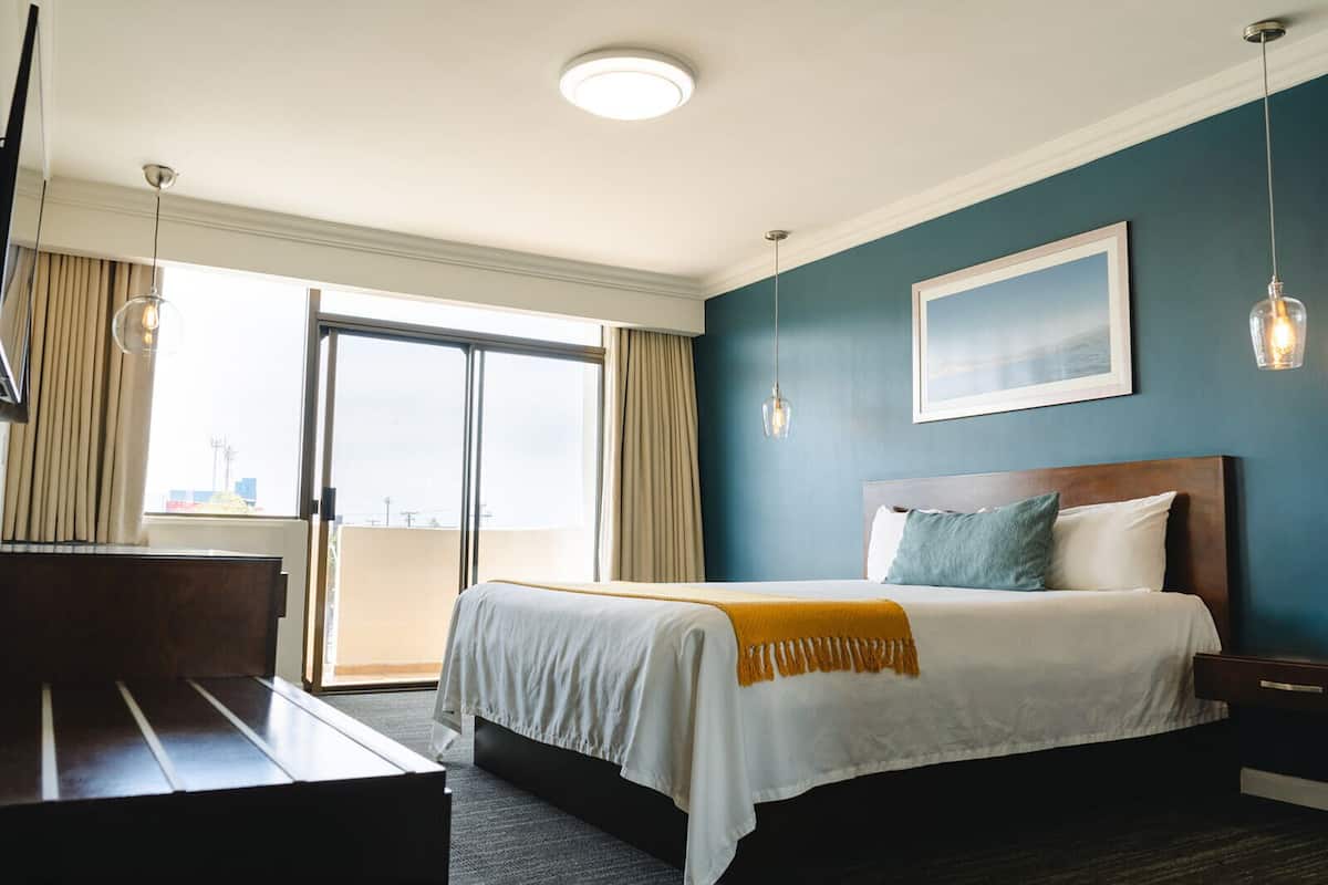 A hotel room with a bed, a desk, a view through a sliding door, and a painting on the wall.